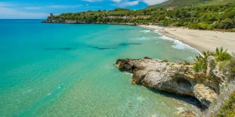 Spiaggia dorata e mare cristallino nel Cilento, con una torre antica su una scogliera e colline verdi sullo sfondo.