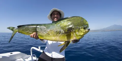 Un pescatore sorridente mostra un grande lampuga, un esempio di cosa si pesca in Sicilia, con il mare blu e una costa all'orizzonte.
