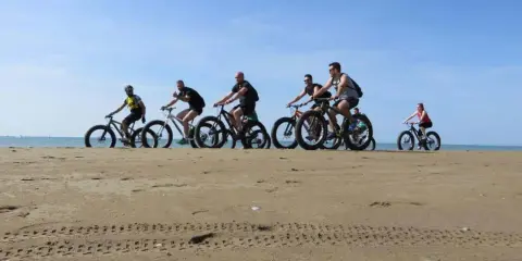 Gruppo di ciclisti su fat bike lungo la ciclabile di Caorle, sulla sabbia dorata con il mare sullo sfondo.