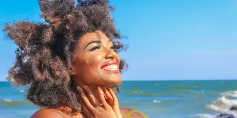 Donna con capelli afro e trucco scintillante si gode il sole in spiaggia. Sembra che ci si abbronza con il trucco, creando un look festoso.