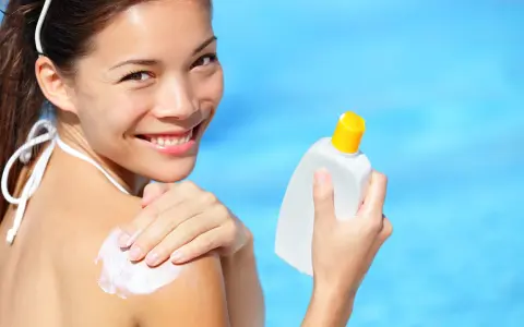 Donna sorridente applica crema solare sulla spalla, con una bottiglia in mano. Scopri che protezione solare usare per la tua pelle.