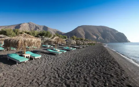 Santorini spiagge di sabbia nera con lettini e ombrelloni di paglia, montagne sullo sfondo e mare calmo.