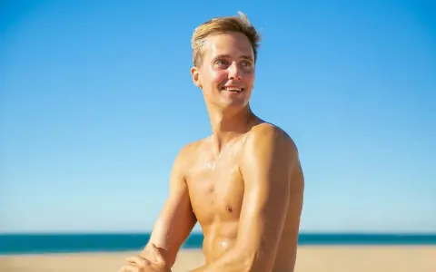 Giovane uomo sorridente sulla spiaggia, con la pelle dorata come se con l'autoabbronzante potesse prendere il sole senza rischi.
