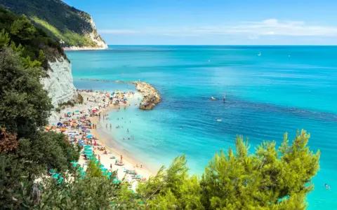 Spiaggia affollata con ombrelloni colorati e gente che nuota nel mare cristallino dell'Abruzzo. Scogliera bianca e vegetazione rigogliosa incorniciano questo angolo di paradiso.