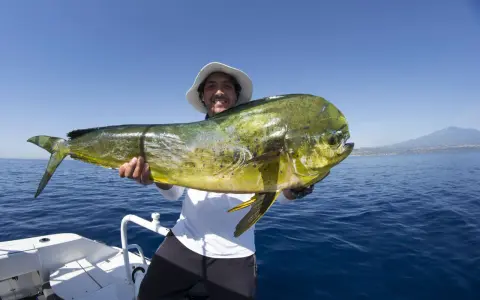 Un pescatore sorridente mostra un grande lampuga, un esempio di cosa si pesca in Sicilia, con il mare blu e una costa all'orizzonte.
