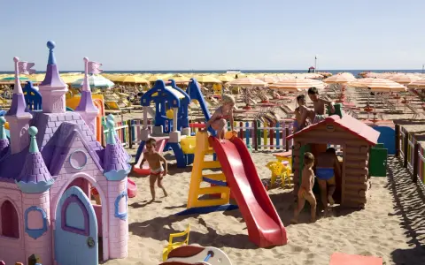 Bambini giocano in un'area giochi sulla spiaggia di Rimini, tra castelli rosa, scivoli e casette di legno. Perfetto per scoprire cosa fare a Rimini con i bambini!
