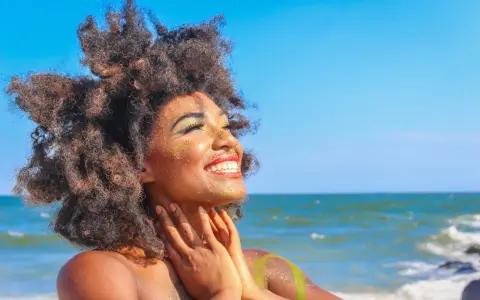 Donna con capelli afro e trucco scintillante si gode il sole in spiaggia. Sembra che ci si abbronza con il trucco, creando un look festoso.