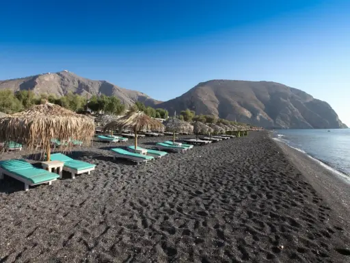 Santorini spiagge di sabbia nera con lettini e ombrelloni di paglia, montagne sullo sfondo e mare calmo.