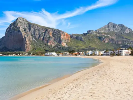 Spiaggia dorata e mare cristallino, un angolo di paradiso in Sicilia dove andare al mare. Montagne imponenti sullo sfondo.