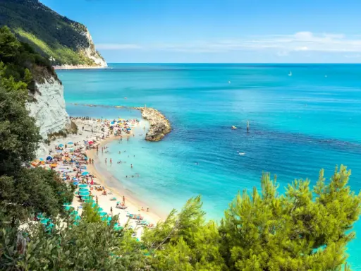Spiaggia affollata con ombrelloni colorati e gente che nuota nel mare cristallino dell'Abruzzo. Scogliera bianca e vegetazione rigogliosa incorniciano questo angolo di paradiso.