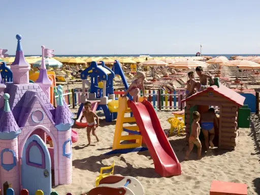 Bambini giocano in un'area giochi sulla spiaggia di Rimini, tra castelli rosa, scivoli e casette di legno. Perfetto per scoprire cosa fare a Rimini con i bambini!