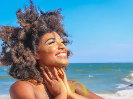 Donna con capelli afro e trucco scintillante si gode il sole in spiaggia. Sembra che ci si abbronza con il trucco, creando un look festoso.