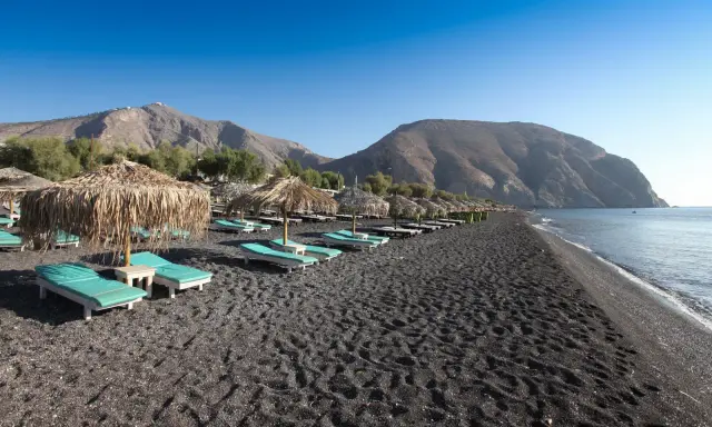 Santorini spiagge di sabbia nera con lettini e ombrelloni di paglia, montagne sullo sfondo e mare calmo.