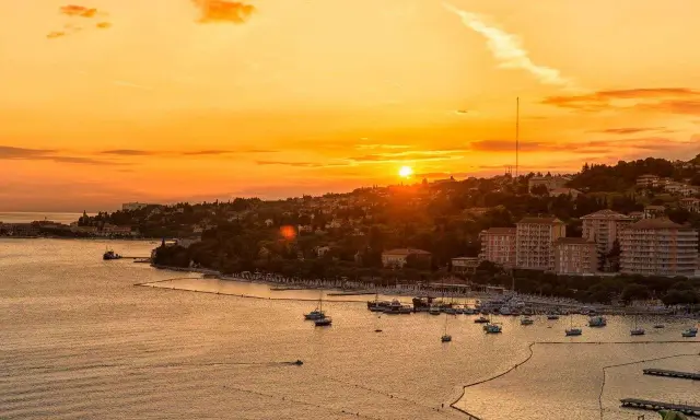 Tramonto dorato sul mare in Slovenia, con un porto pieno di barche e una costa verdeggiante. Immagina i bambini che giocano felici sulla spiaggia.