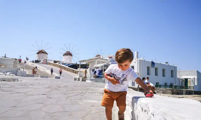 Un bambino gioca con una macchinina rossa a Mykonos, con i mulini a vento sullo sfondo. Vacanze a Mykonos con bambini.