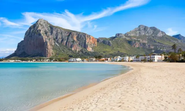 Spiaggia dorata e mare cristallino, un angolo di paradiso in Sicilia dove andare al mare. Montagne imponenti sullo sfondo.
