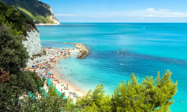 Spiaggia affollata con ombrelloni colorati e gente che nuota nel mare cristallino dell'Abruzzo. Scogliera bianca e vegetazione rigogliosa incorniciano questo angolo di paradiso.