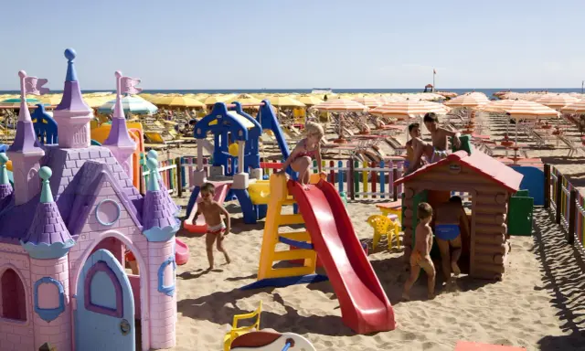 Bambini giocano in un'area giochi sulla spiaggia di Rimini, tra castelli rosa, scivoli e casette di legno. Perfetto per scoprire cosa fare a Rimini con i bambini!