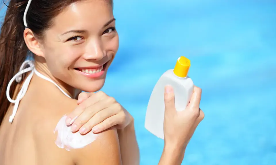 Donna sorridente applica crema solare sulla spalla, con una bottiglia in mano. Scopri che protezione solare usare per la tua pelle.