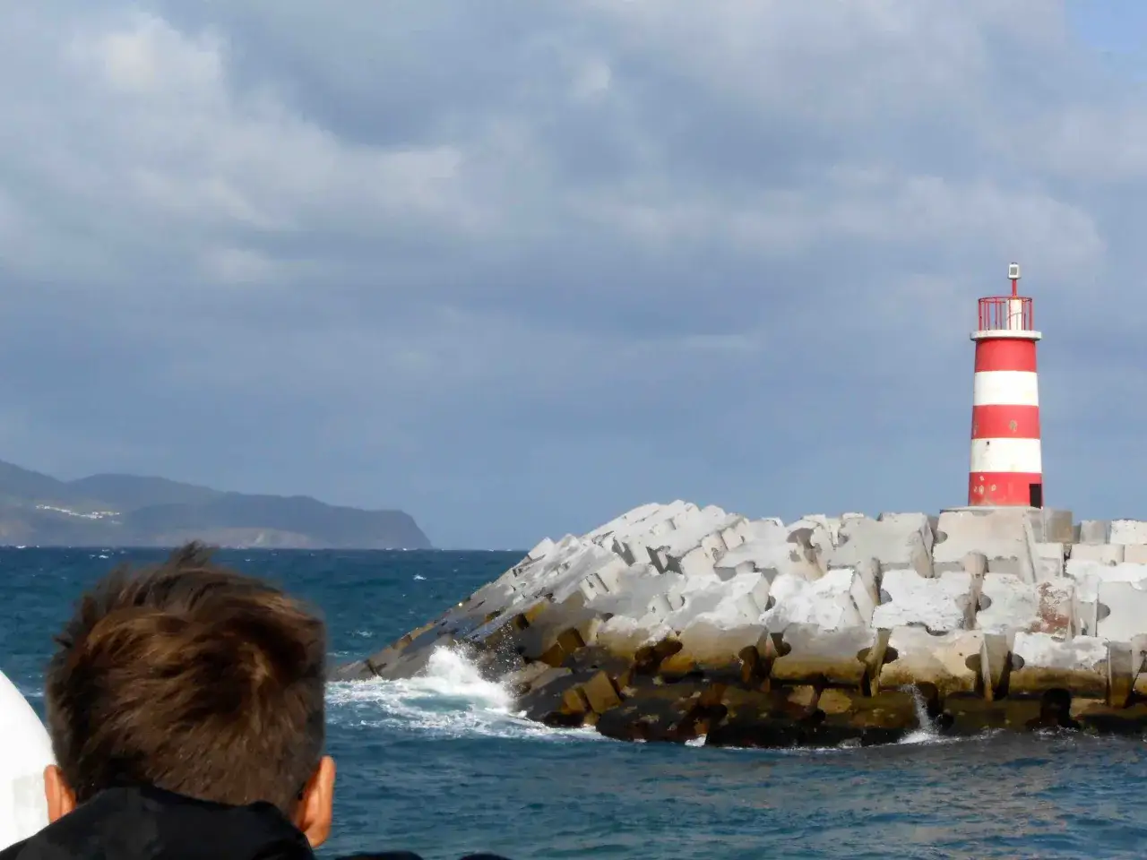 Un bambino guarda un faro a strisce rosse e bianche sulle Azzorre, con onde che si infrangono su blocchi di cemento.