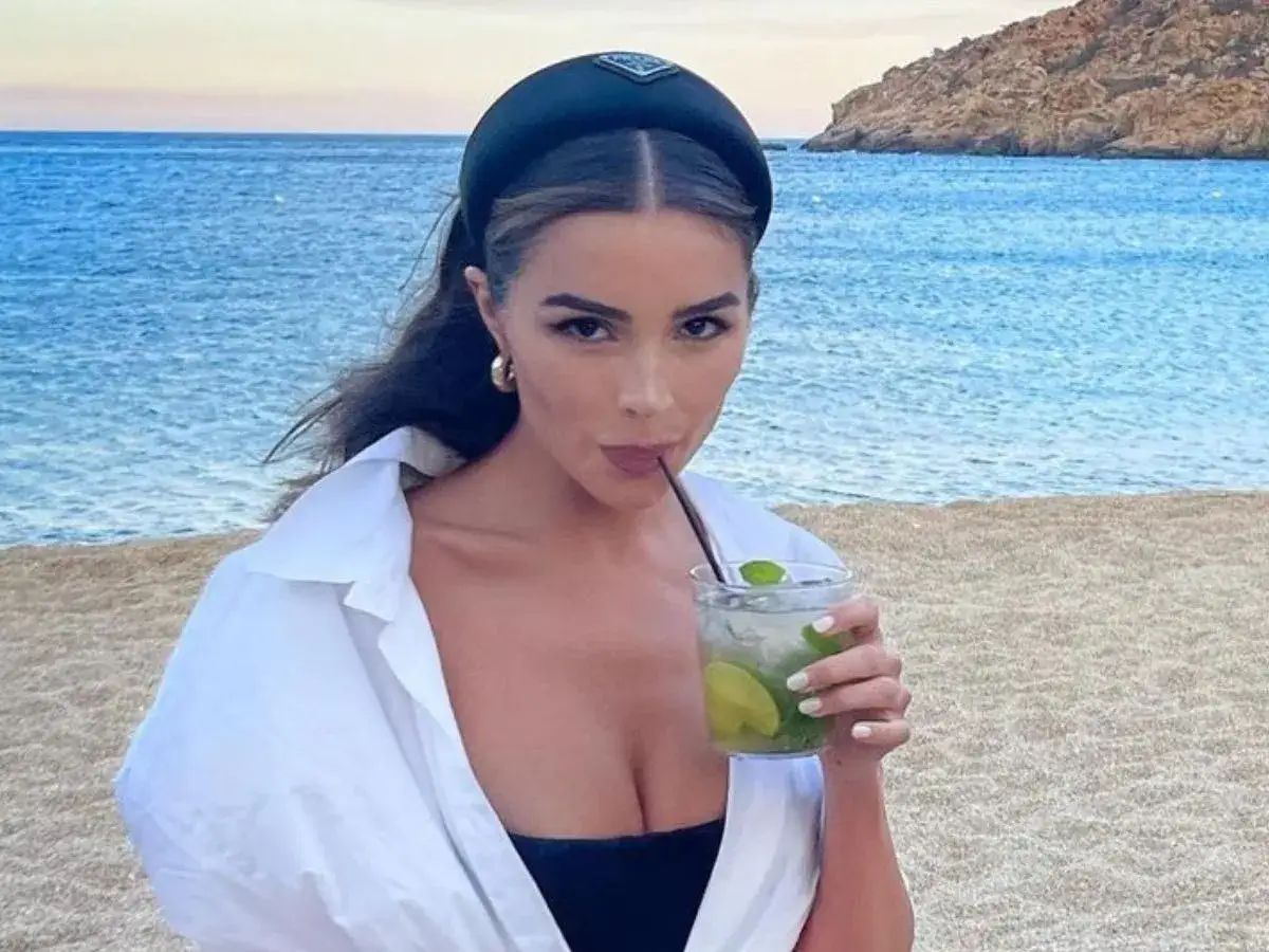 Donna con cerchietto Prada beve un mojito sulla spiaggia. Sembra che ci si abbronza con il trucco, con un look da vacanza perfetto.
