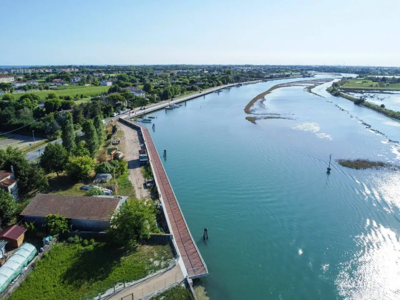 La ciclabile Cavallino Treporti si estende lungo un canale azzurro, con case e vegetazione a sinistra e un paesaggio lagunare a destra.