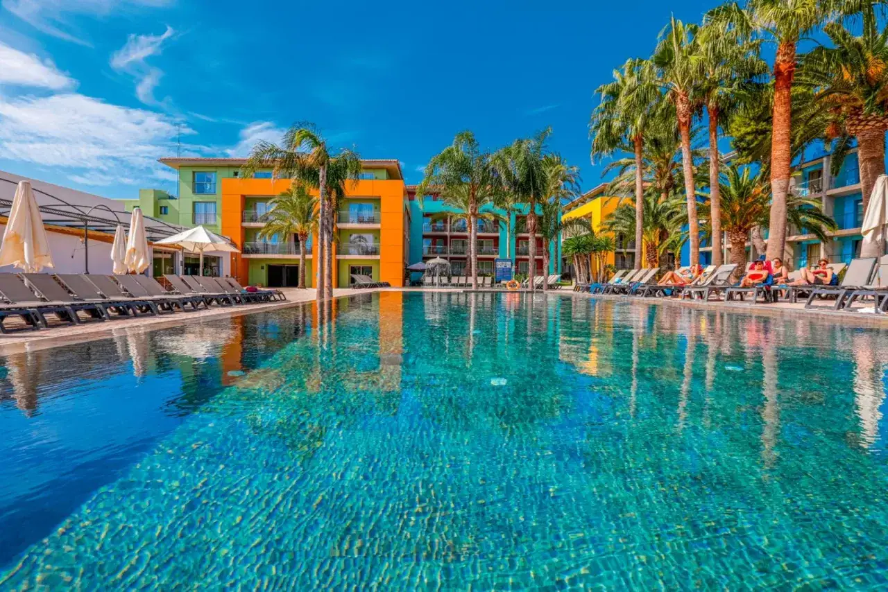 Piscina scintillante e palme rigogliose al hotel Occidental Menorca, con edifici colorati sullo sfondo e persone che si rilassano sui lettini.