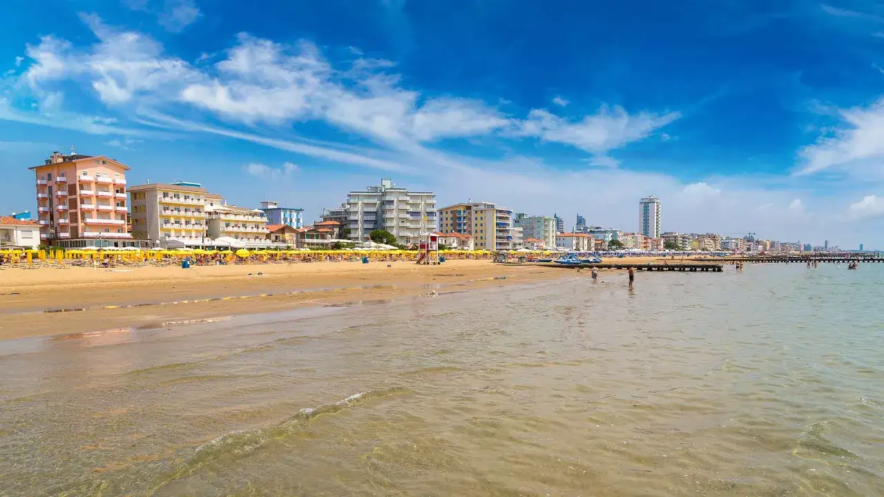 Spiaggia affollata con ombrelloni gialli e hotel sul lungomare, bagnanti in acqua e un molo nel mare veneto sotto un cielo azzurro con nuvole bianche.