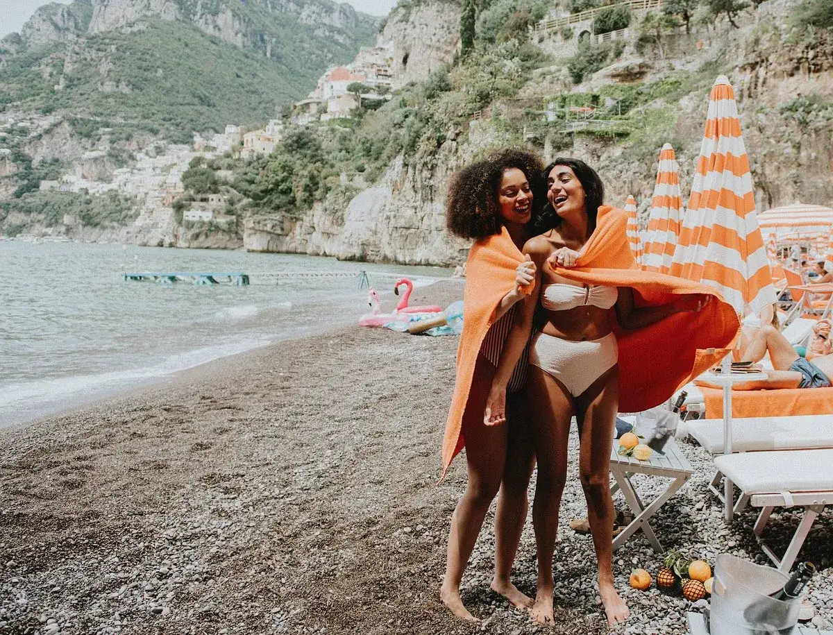Due amiche ridono sulla spiaggia, avvolte in un telo arancione. Un'estate a Cipro con bambini, tra mare e divertimento.