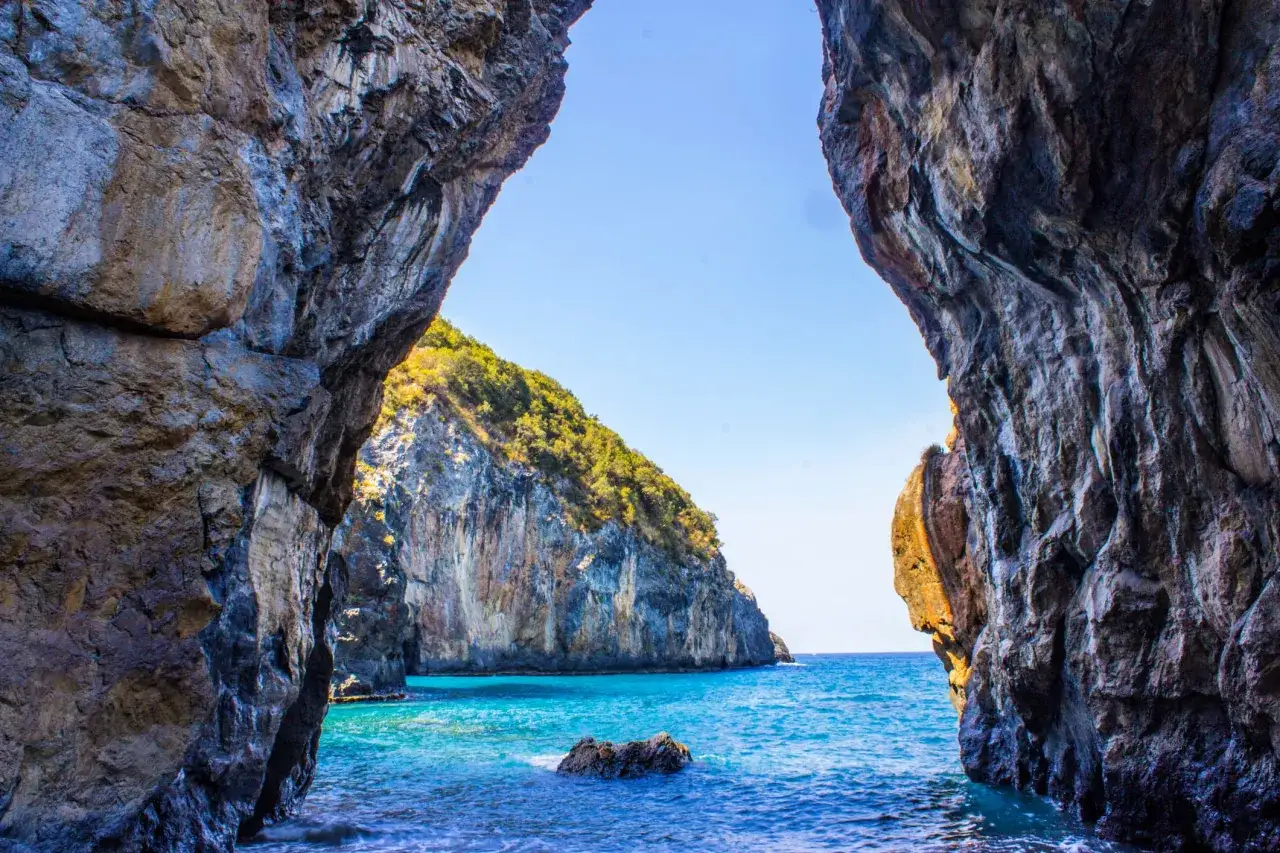 Vista dal mare attraverso un arco naturale di roccia. La scogliera verdeggiante si affaccia su acque turchesi cristalline.