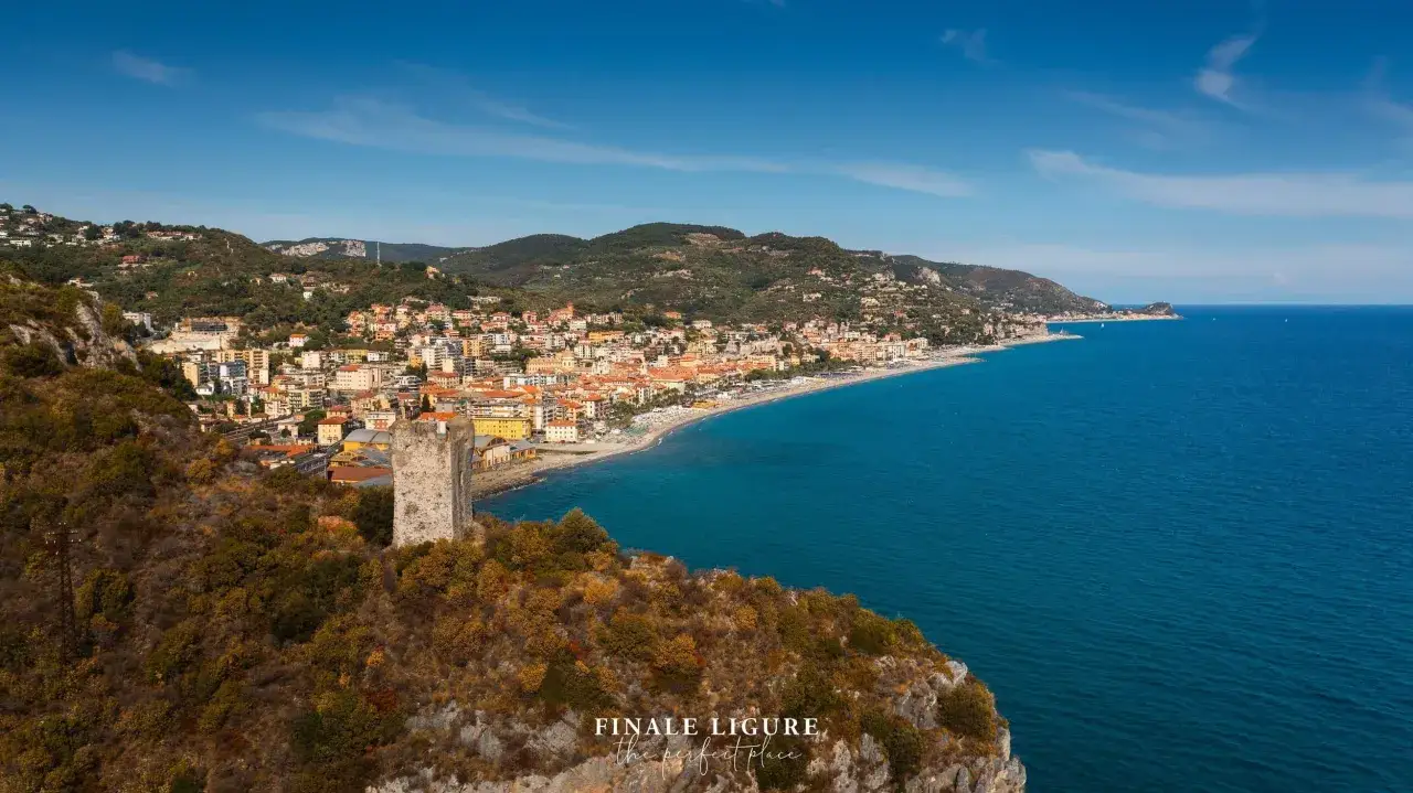 Vista panoramica della spiaggia di Finale Ligure con il mare azzurro, le case colorate e una torre antica su una collina rocciosa.