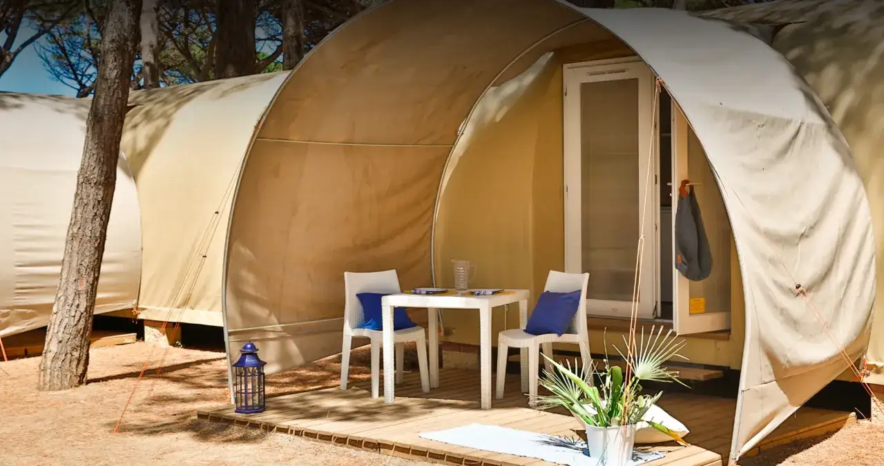 Tavolino e sedie con cuscini blu davanti a una tenda del Gitavillage Il Gabbiano, immersa tra i pini.