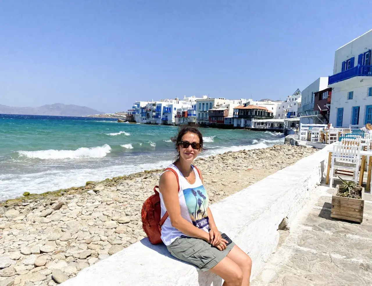 Donna sorridente a Mykonos, con le case bianche e blu e il mare turchese sullo sfondo. Un luogo perfetto per una vacanza con bambini.