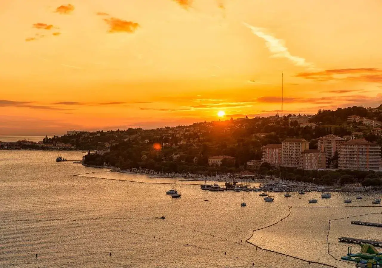Tramonto dorato sul mare in Slovenia, con un porto pieno di barche e una costa verdeggiante. Immagina i bambini che giocano felici sulla spiaggia.