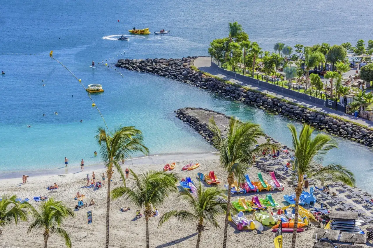 Spiaggia a Gran Canaria con bambini che giocano, pedalò colorati e palme. L'acqua turchese invita a nuotare.