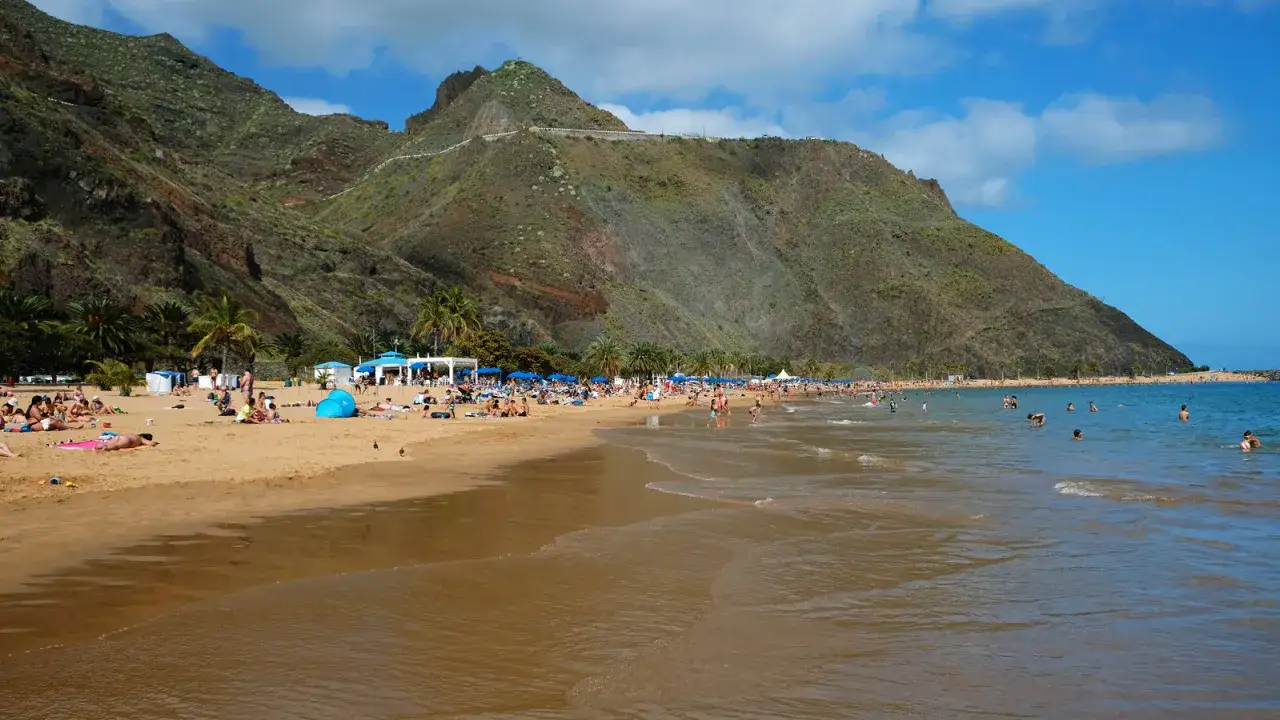 Spiaggia affollata a Tenerife con bambini che giocano nell'acqua e famiglie che si rilassano sulla sabbia dorata, sotto imponenti montagne verdi.