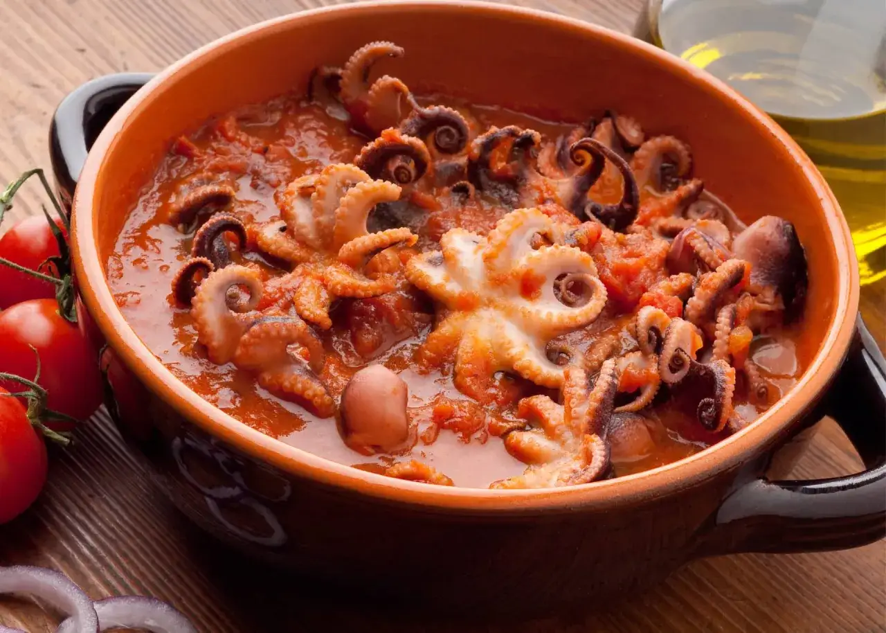 Polpo alla luciana in un tegame di terracotta, con pomodoro e contorni di pomodorini e cipolla rossa.