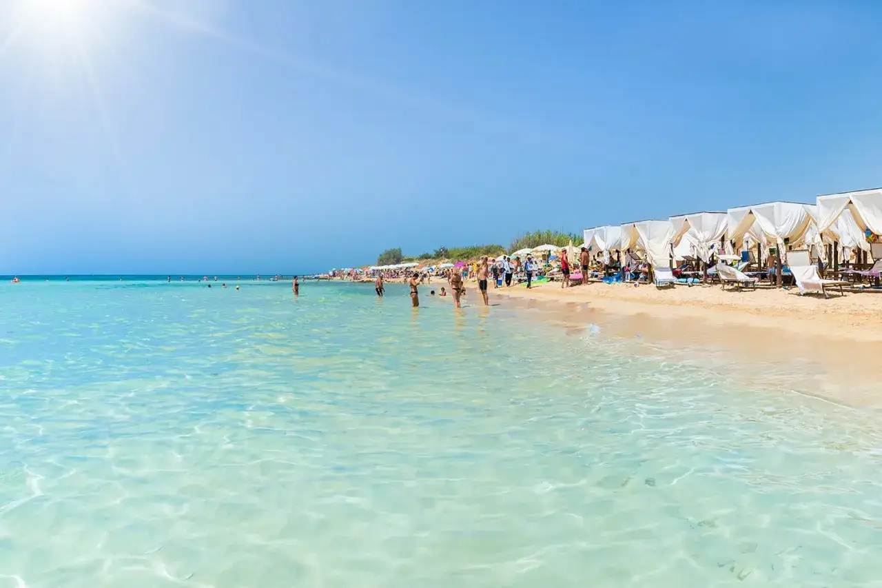 Spiaggia in Puglia con bambini che giocano nell'acqua cristallina e famiglie che si rilassano sotto i gazebo bianchi.