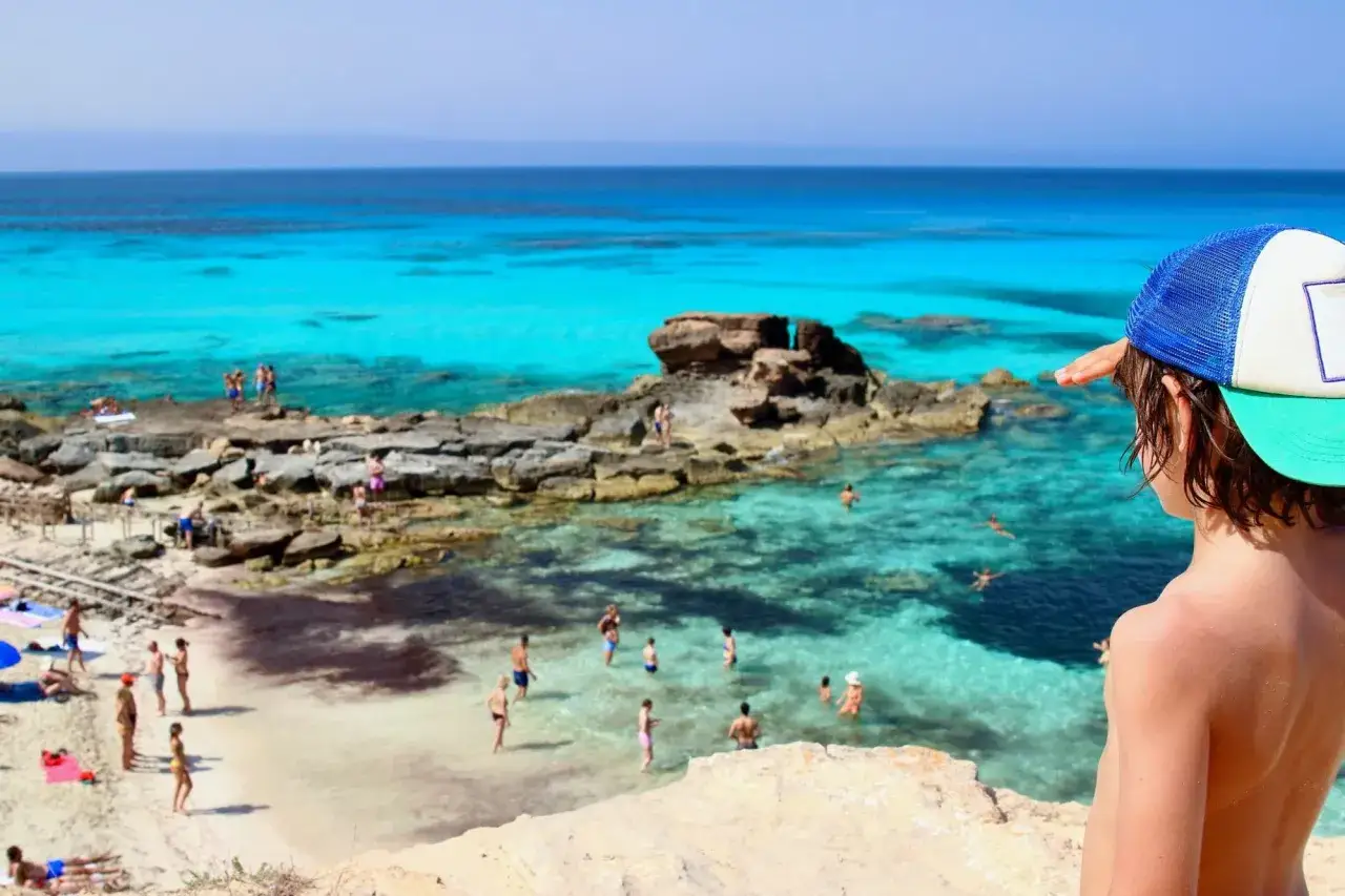 Bambino con cappellino guarda il mare cristallino di Formentera, dove bagnanti si godono la spiaggia e le acque turchesi. Vacanze a Formentera con bambini.