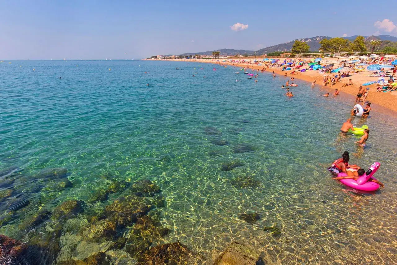 Spiaggia affollata a Santa Susanna, Spagna, con persone che nuotano e si rilassano in acqua cristallina.