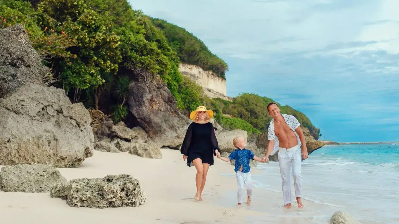 Famiglia felice in vacanza a Bali con bambini, passeggiando sulla spiaggia con rocce e vegetazione lussureggiante.