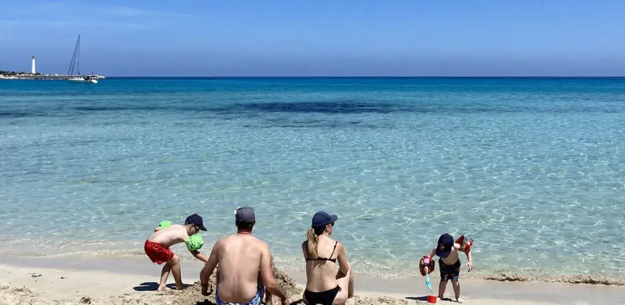 Famiglia felice a San Vito Lo Capo con bambini che giocano sulla sabbia, mare cristallino e un veliero all'orizzonte.