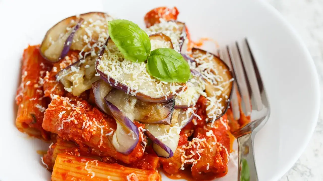 Un piatto di pasta alla norma con melanzane fritte, sugo di pomodoro e ricotta salata grattugiata, guarnito con basilico fresco.