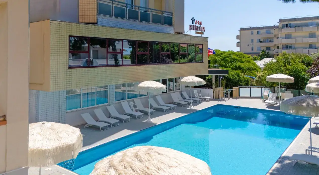 Piscina azzurra e lettini prendisole al hotel Simon Gatteo Mare. Ombrelloni di paglia offrono riparo dal sole.