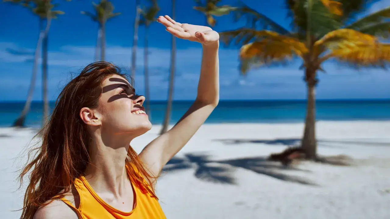 Ragazza con abbronzatura a chiazze sulla spiaggia, si copre il viso dal sole con la mano. Palme e mare sullo sfondo.