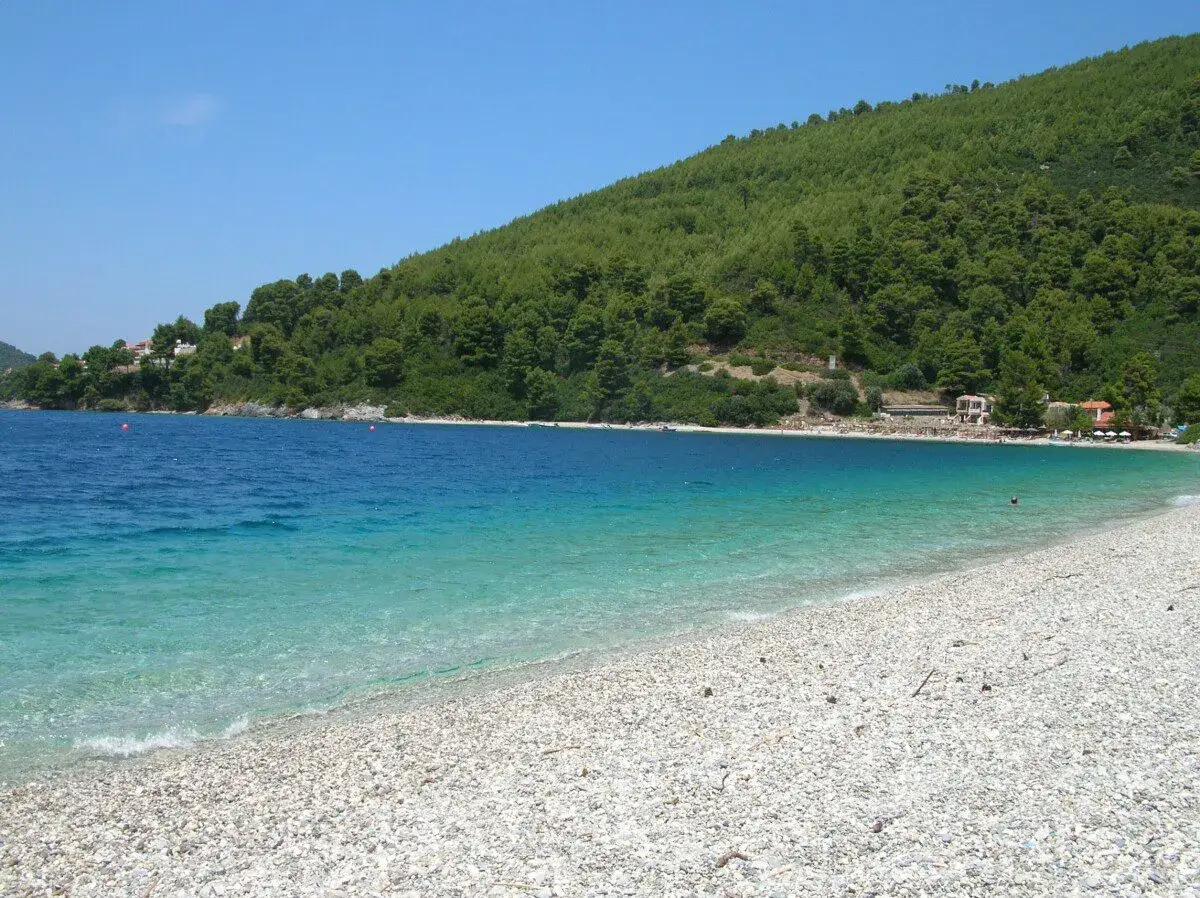 Acque cristalline e una spiaggia di ciottoli bianchi invitano al relax su una delle splendide alonissos spiagge, con una collina verdeggiante a fare da sfondo.