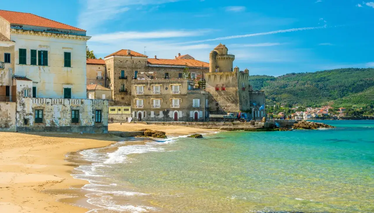 Spiaggia dorata e mare cristallino nel Cilento, con antichi palazzi e un castello che si affacciano sull'acqua turchese.