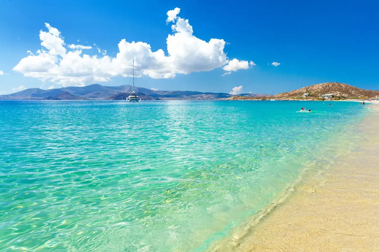 Spiaggia di Naxos con bambini che nuotano nelle acque cristalline, un catamarano all'orizzonte e montagne sullo sfondo.