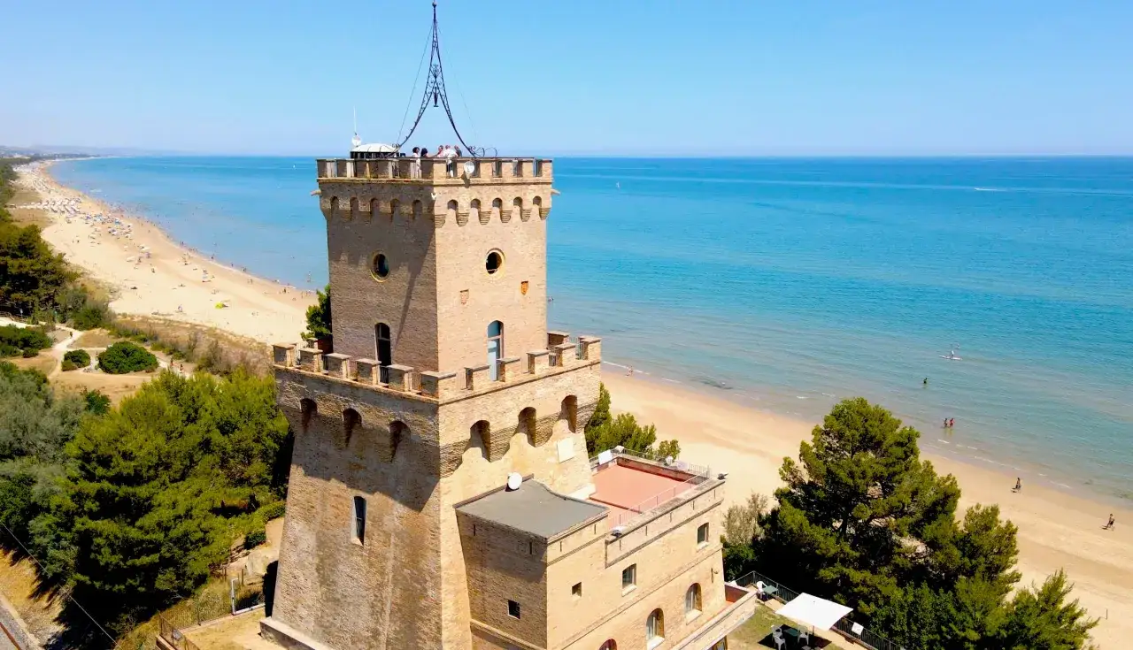 Torre medievale affacciata sul pineto mare, con spiaggia dorata e mare azzurro.