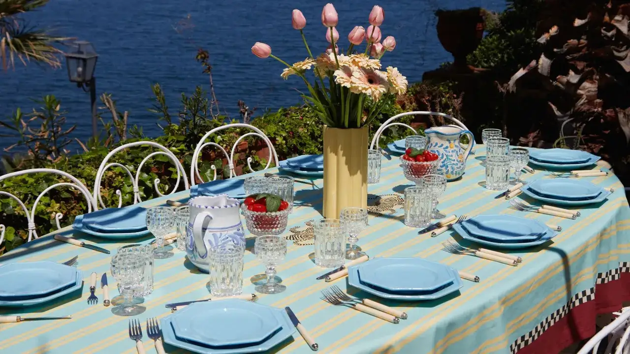 Tavola apparecchiata per Ferragosto con piatti azzurri, fiori e vista mare.